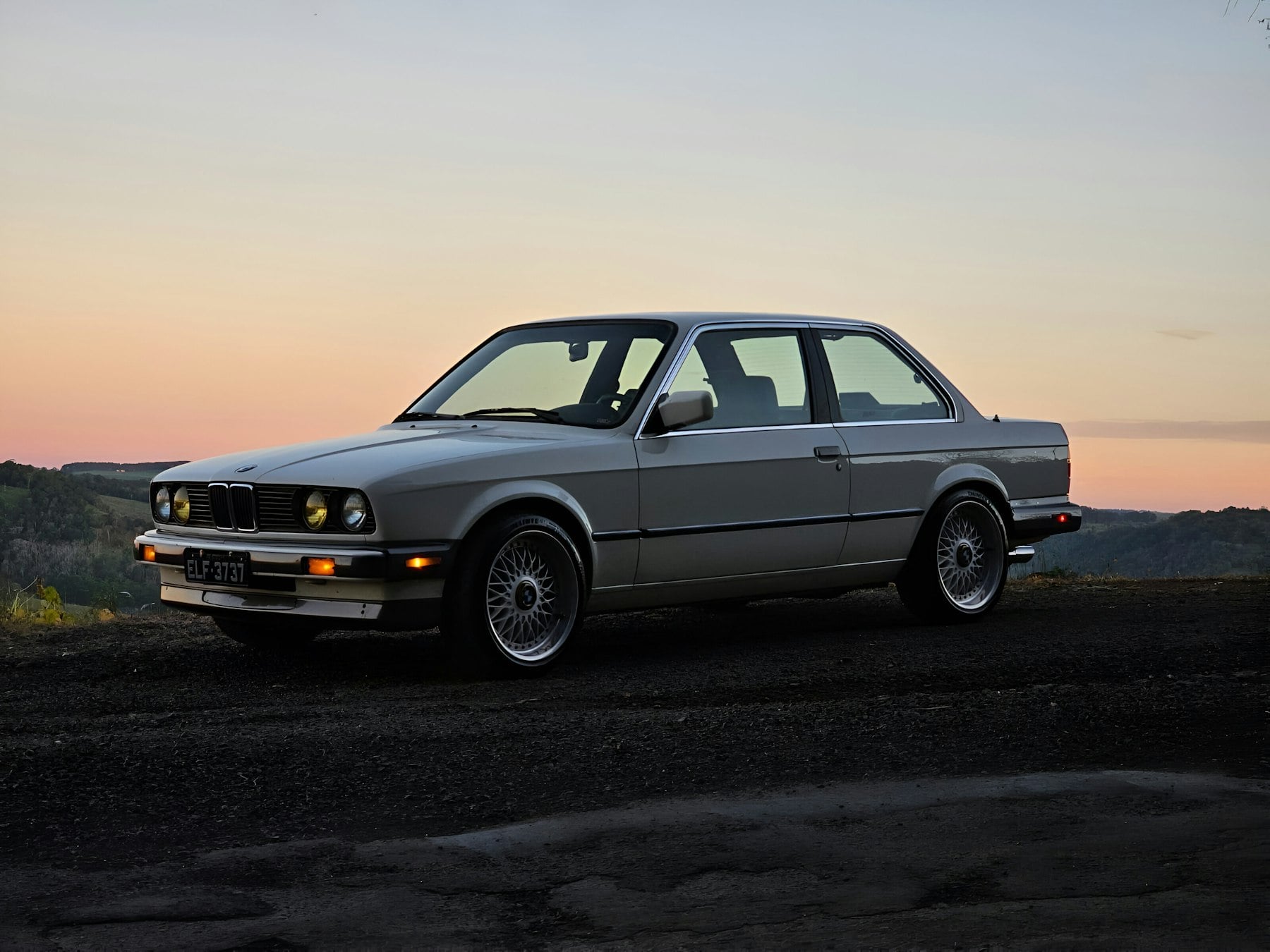 An Alpine White BMW E30 coupe on BBS basketweave wheels, parked at dusk